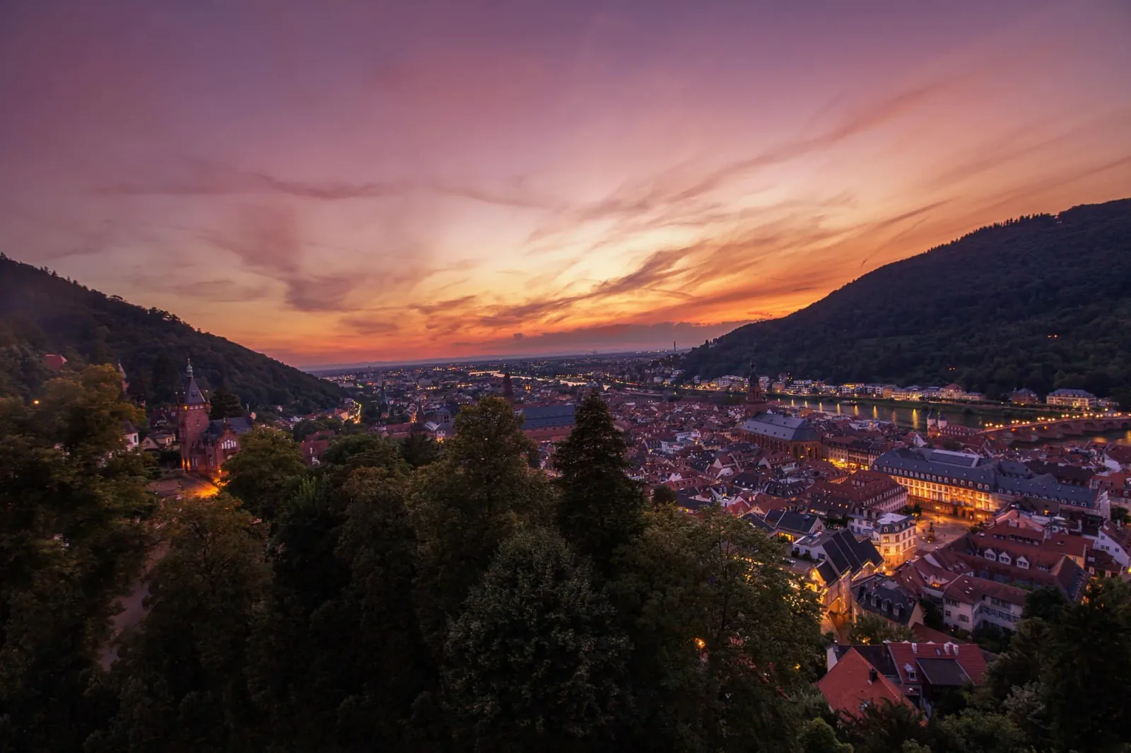 A town in a valley between hills, its lights already on under a violet sky