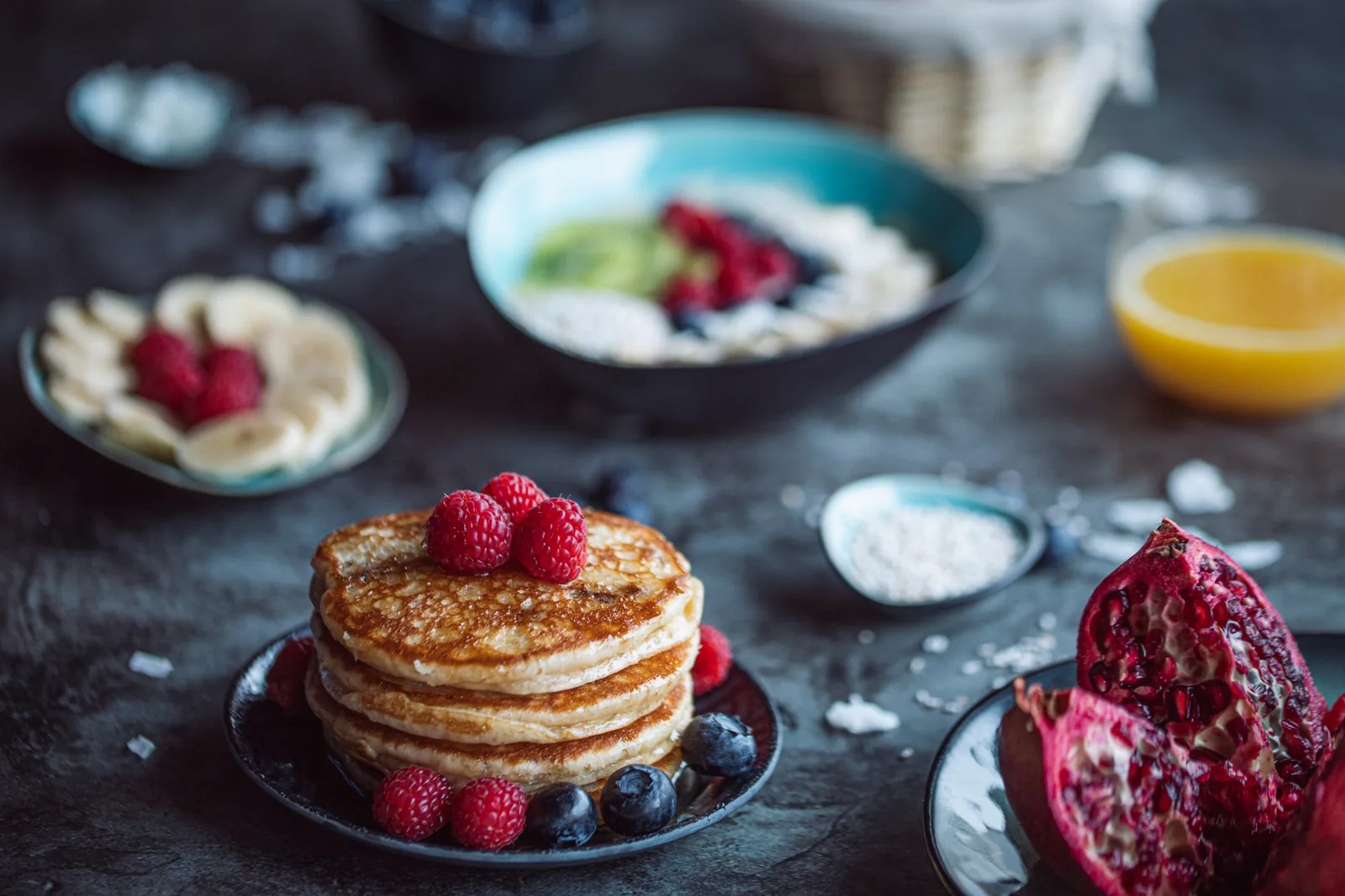 Pancakes with raspberries on dark slate, with a bowl, an orange and a pomegranate behind