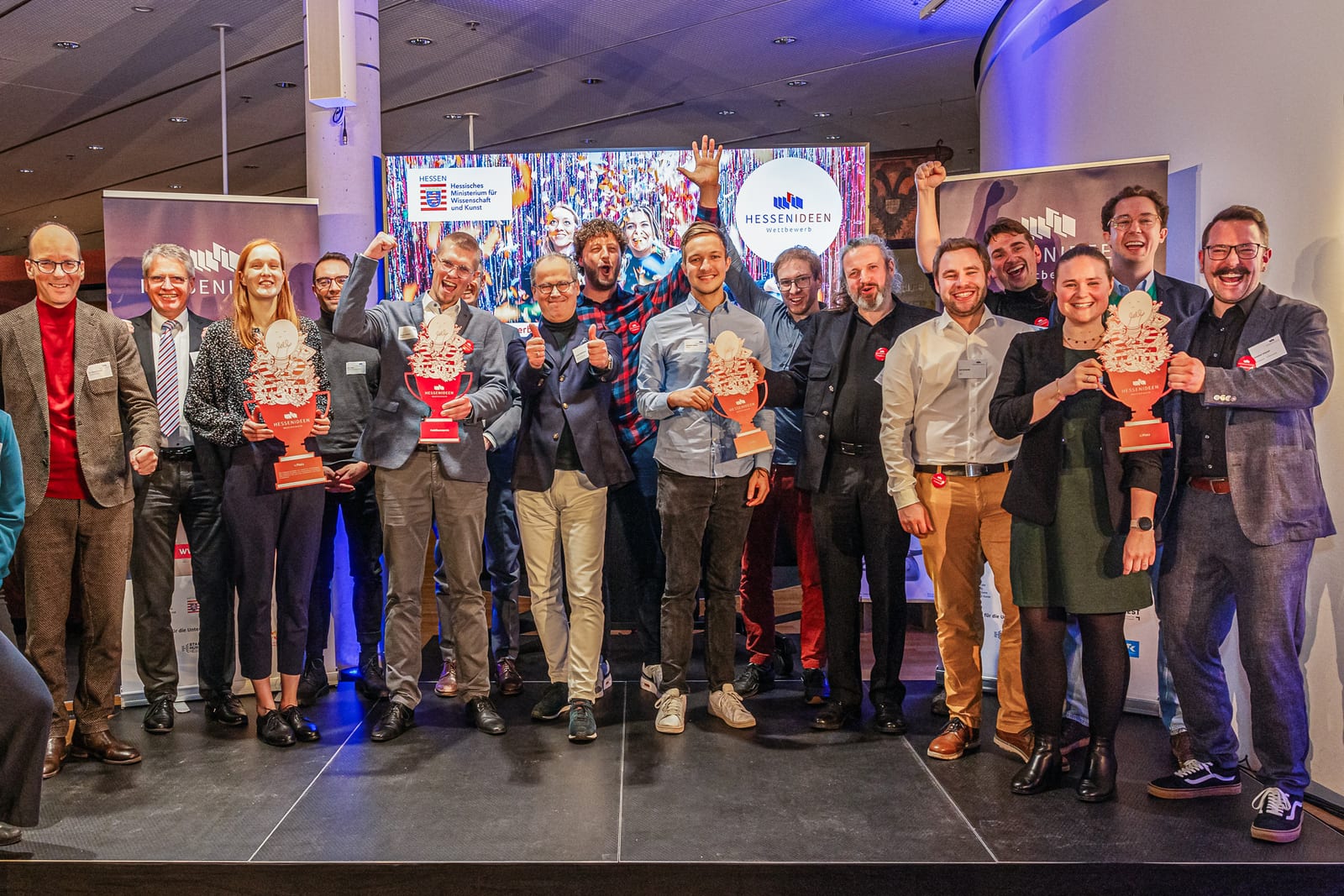 Gruppenbild der Preisträger des Hessen-Ideen-Wettbewerbs auf der Bühne, mehrere halten ihre Trophäen in die Kamera, dahinter die Banner des Wettbewerbs
