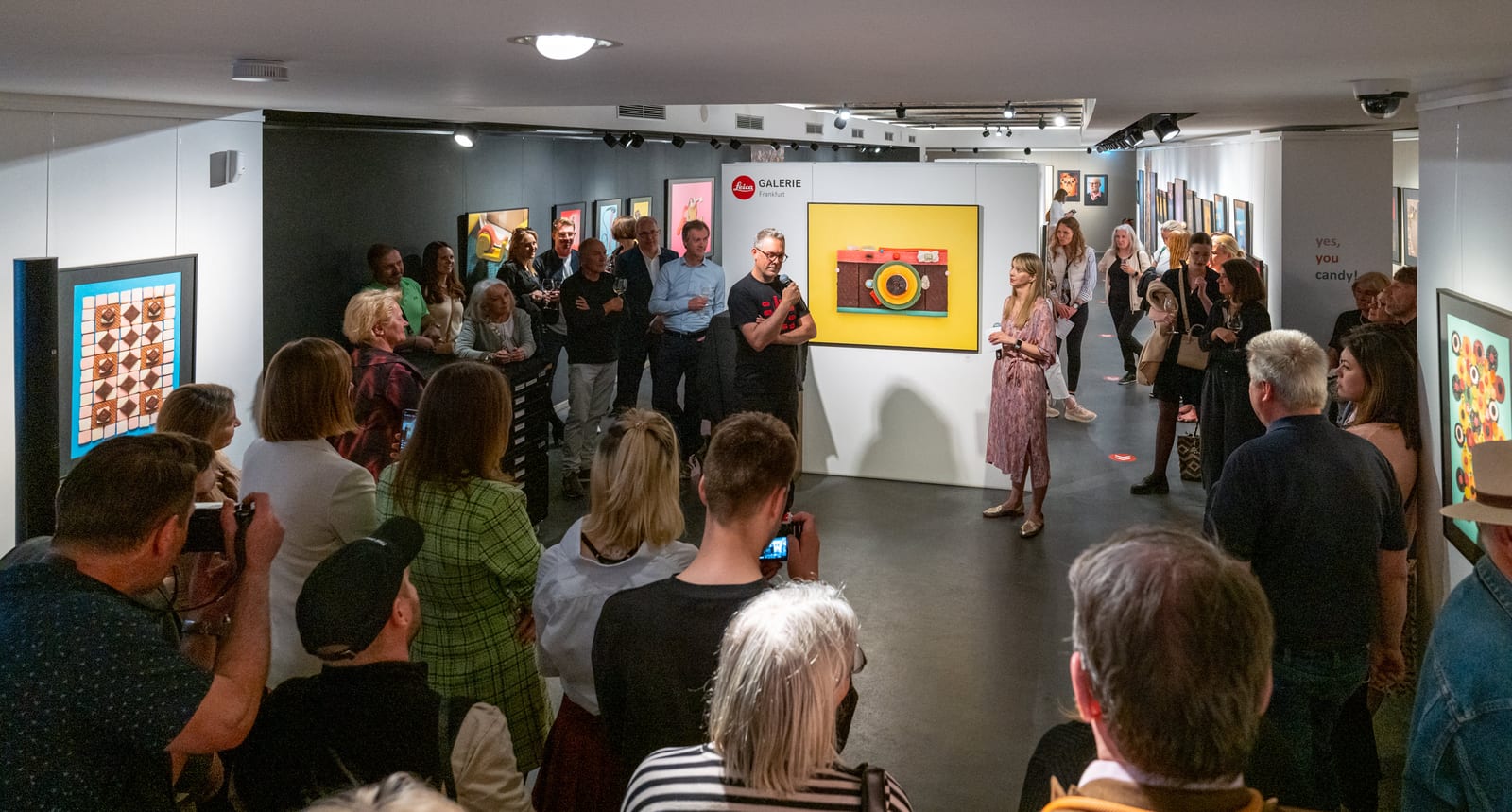 A private view at the Leica Galerie Frankfurt: the artist speaking into a microphone in front of a large print, the audience standing close among the hung works