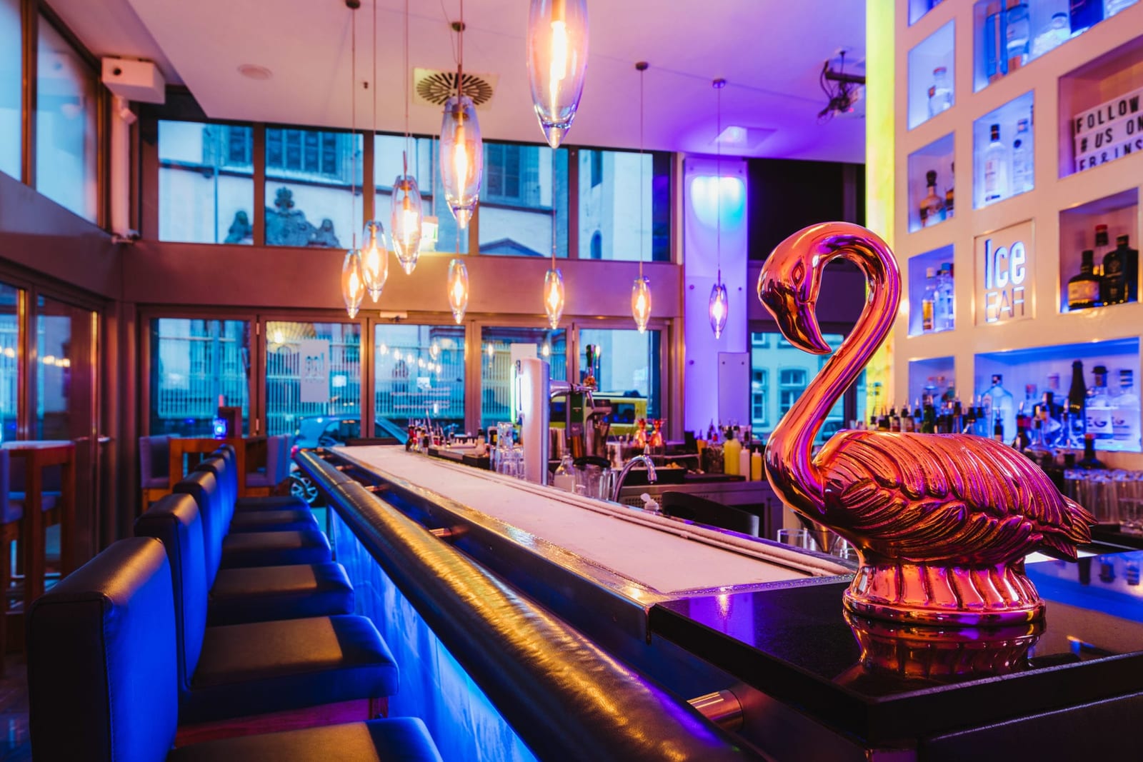 Bar im Hilton Köln bei blau-violettem Licht: lange Theke mit Barhockern, hängende Glühlampen und eine glänzende rosafarbene Flamingofigur im Vordergrund
