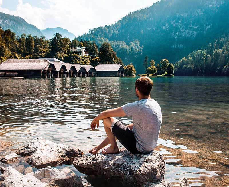 Bergsee mit Bootshäusern, eine Person sitzt am Ufer