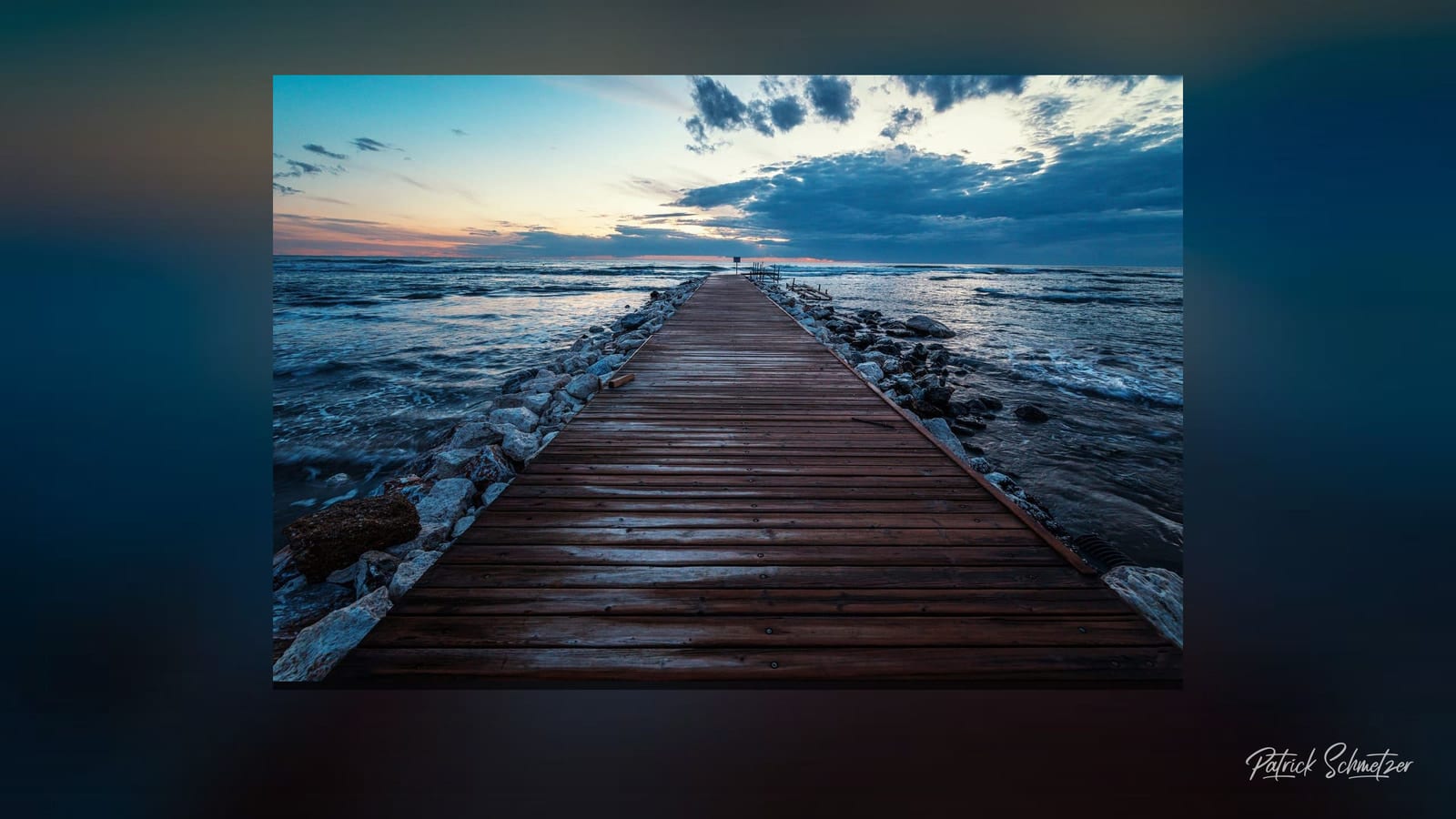 Desktop wallpaper: a wooden jetty running out into the surf on a softly blurred ground, with the signature at the bottom right